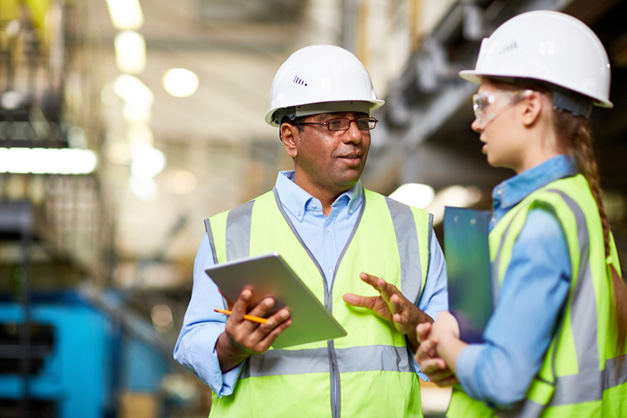 two construction workers in safety gear discussing plans with a tablet and clipboard in an industrial setting